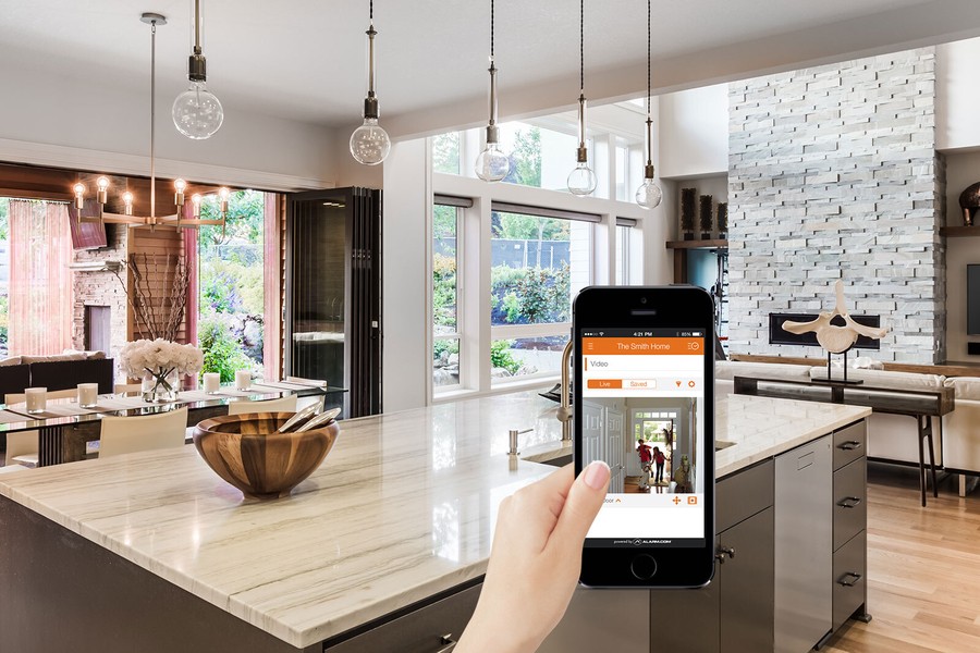 A woman’s hand holding a smartphone with the Alarm.com app in a kitchen area.